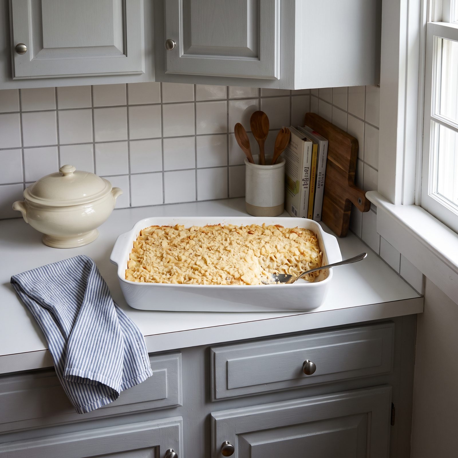 Kitchen counter with a mostly full tuna noodle casserole in a white dish topped with crushed potato chips, serving spoon