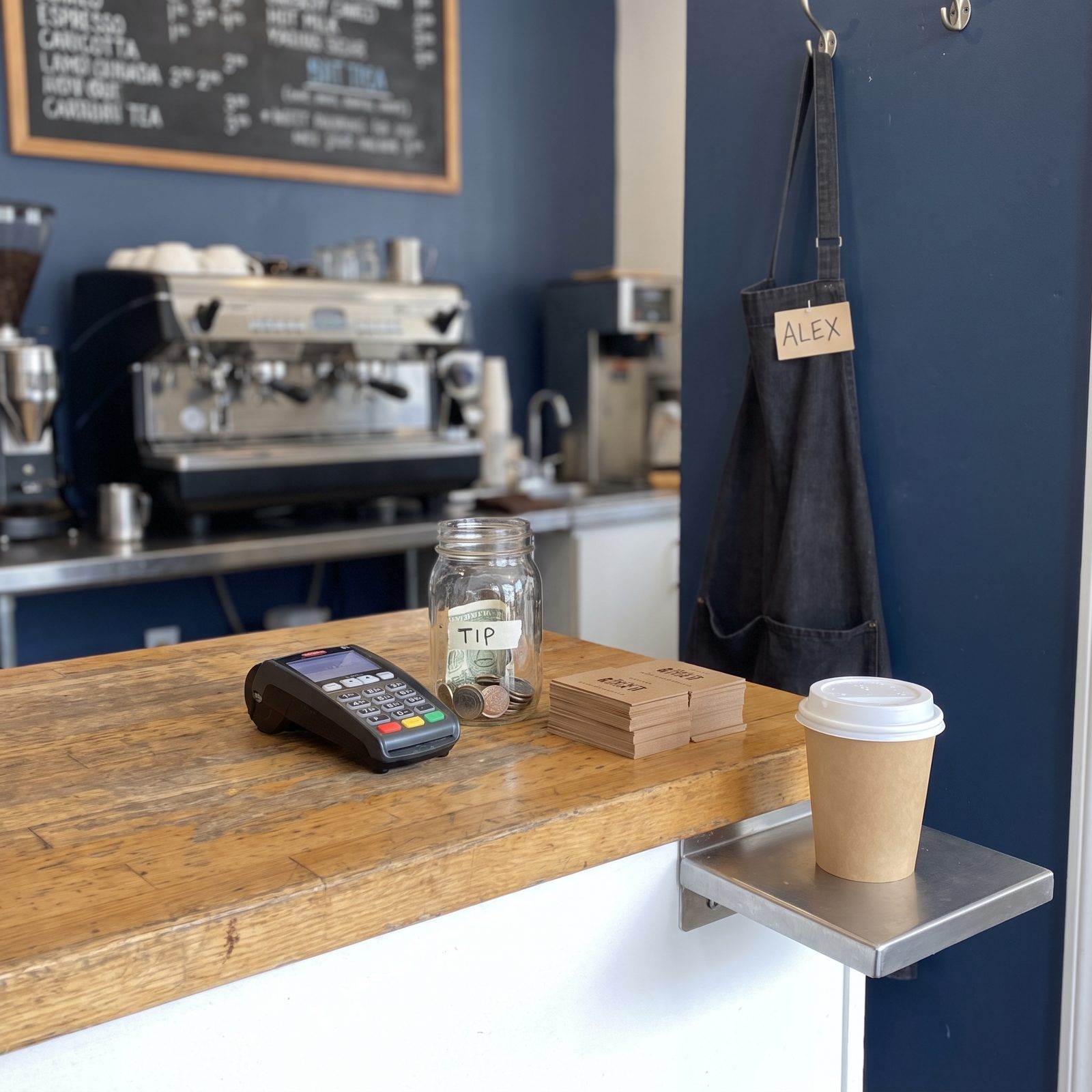 Café order counter with a card reader, tip jar, loyalty cards, and a paper coffee cup on the pickup shelf.