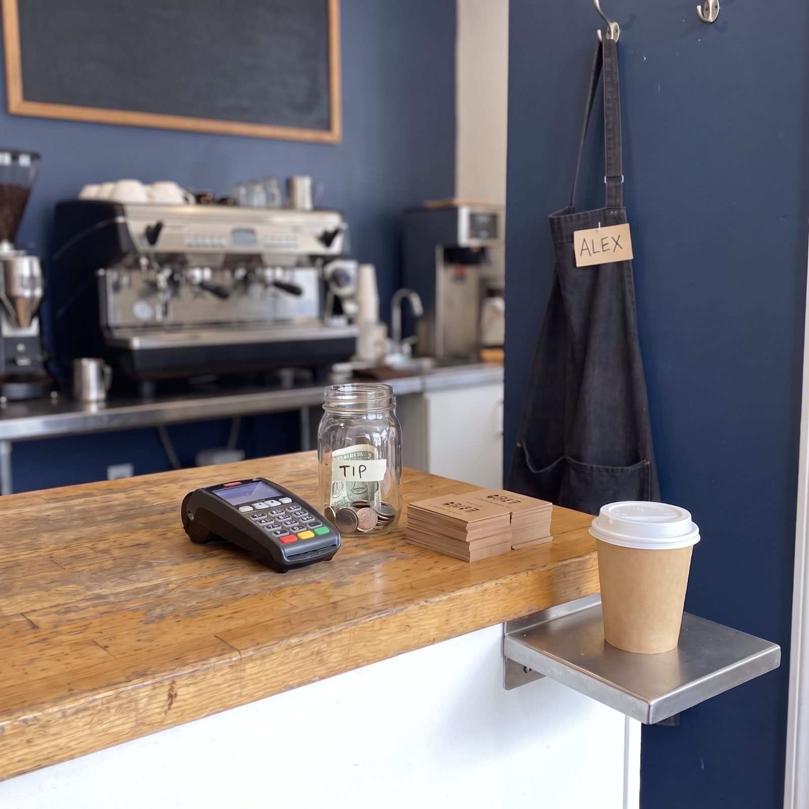 Café order counter with a card reader, tip jar, loyalty cards, and a paper coffee cup on the pickup shelf.